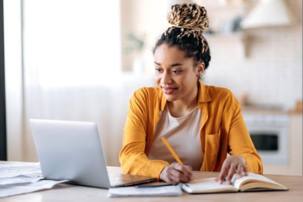 A PhD student writing in a notebook and looking at a laptop..