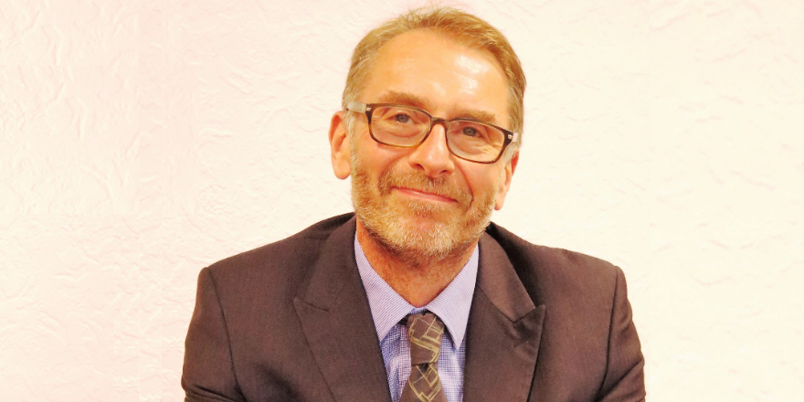 Headshot of man in grey suit jacket with purple tie and glasses.