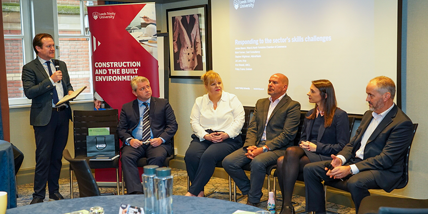 A panel of men and women sitting down in front of a branded Leeds Trinity University banner and having a conversation..