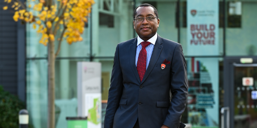 Professor Charles Egbu stands in front of Leeds Trinity University atrium in suit with blue shirt and red tie.