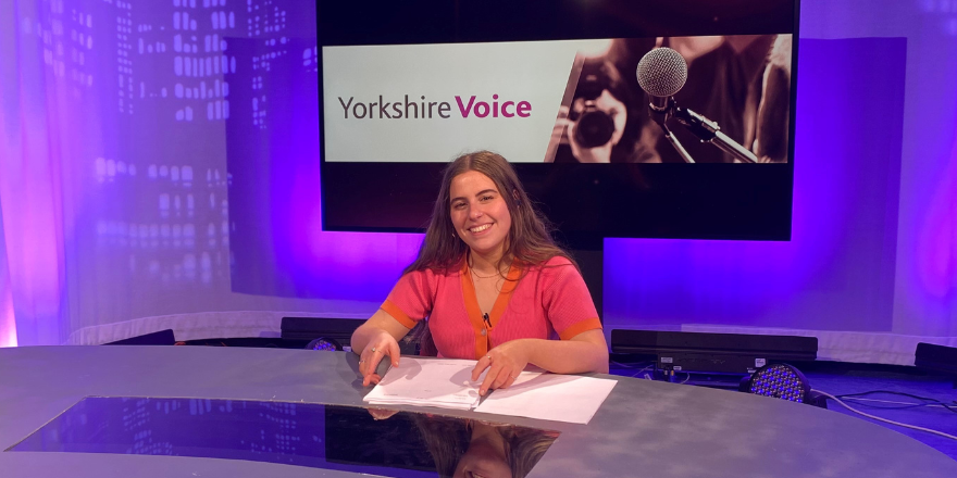 Female student sits at news desk smiling.