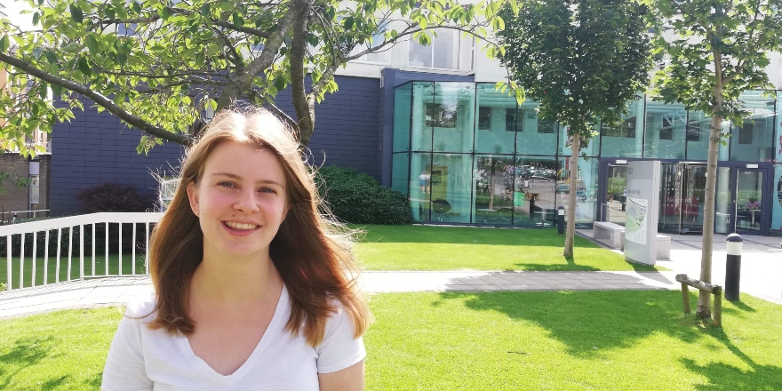 Girl with white top and shoulder length brown hair stands outside campus entrance.