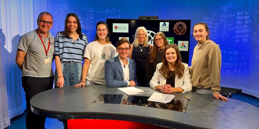 Group of male and female students and tutors stand behind red desk in front of television screen.