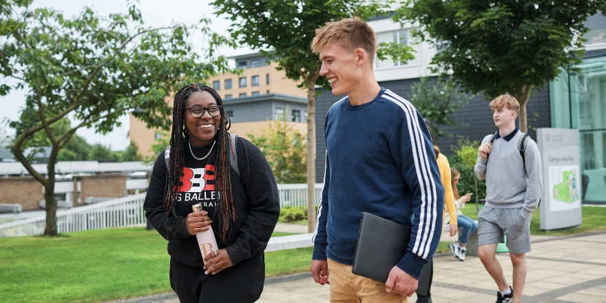 Two students walking and talking at the entrance to campus, with other students and trees in the background.