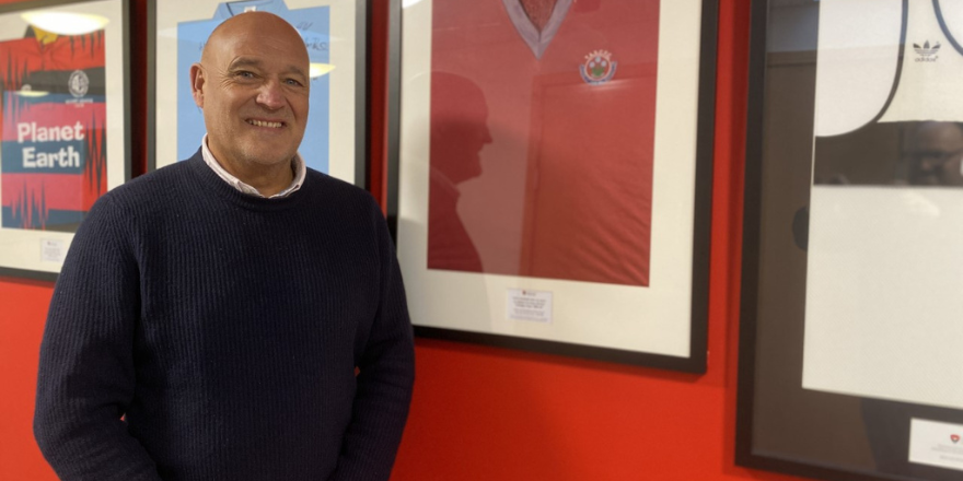 Older man stands in front of framed shirt on wall.