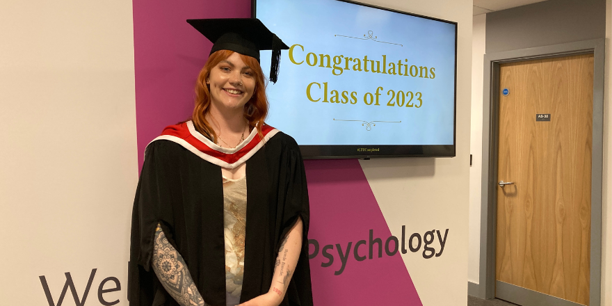 A girl with ginger hair wearing a black cap and gown poses in front of a screen that says, Congratulations Class of 2023.