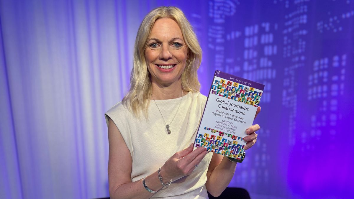 A woman with blonde hair wearing a white dress holds a book.