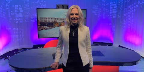 Blonde woman stands in front of broadcast desk and television screen.