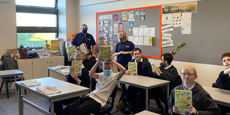 Children in classroom sat at desks holding science books with adult female and adult male stood at back.