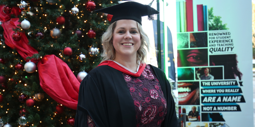 Blonde female in graduation cap stands in front of Christmas tree.