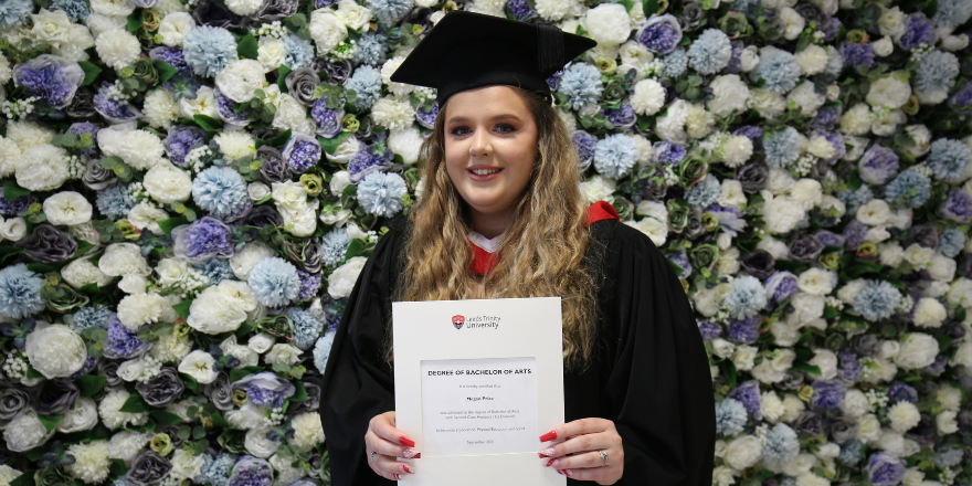 Blonde girl stands in front of flower wall with gown and graduation cap on holding degree certificate.