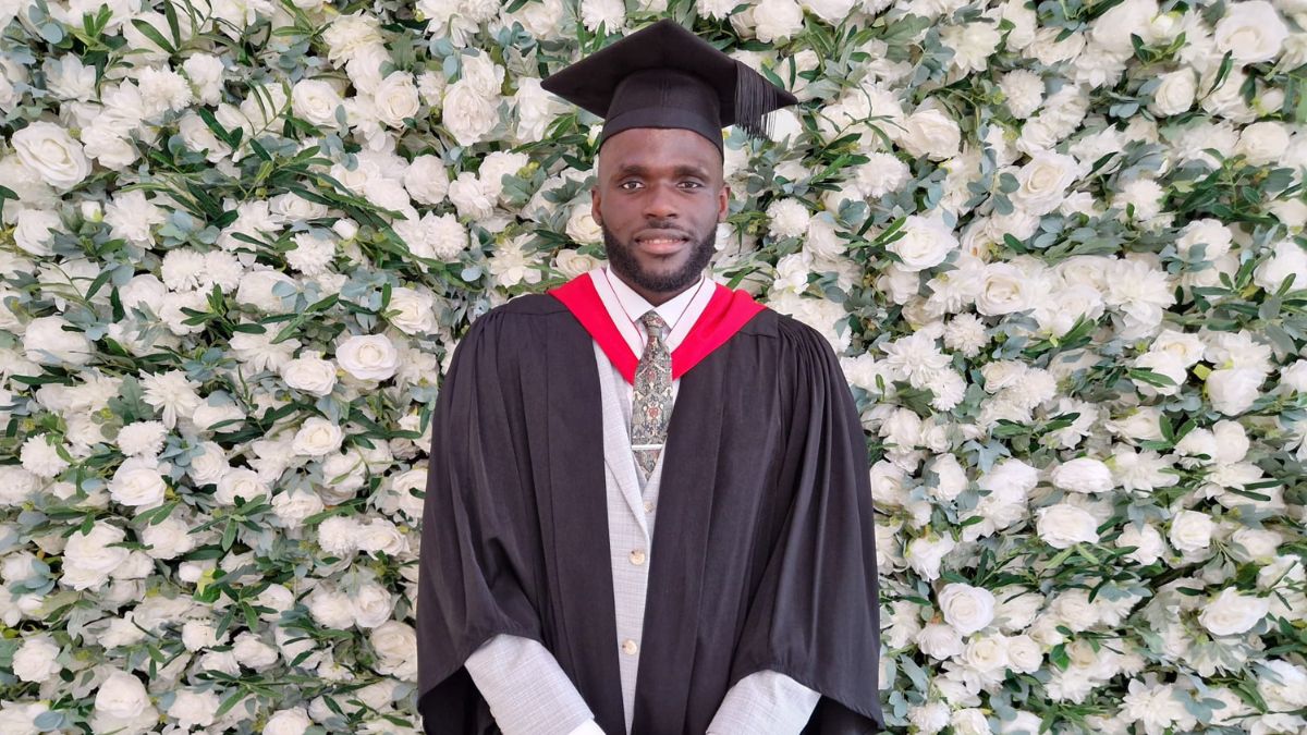 A student wearing a graduation cap and gown stands in front of a flower wall.