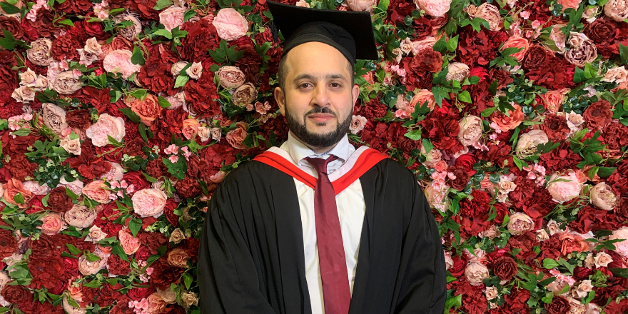 Male in graduation cap and gown stands in front of flower wall.