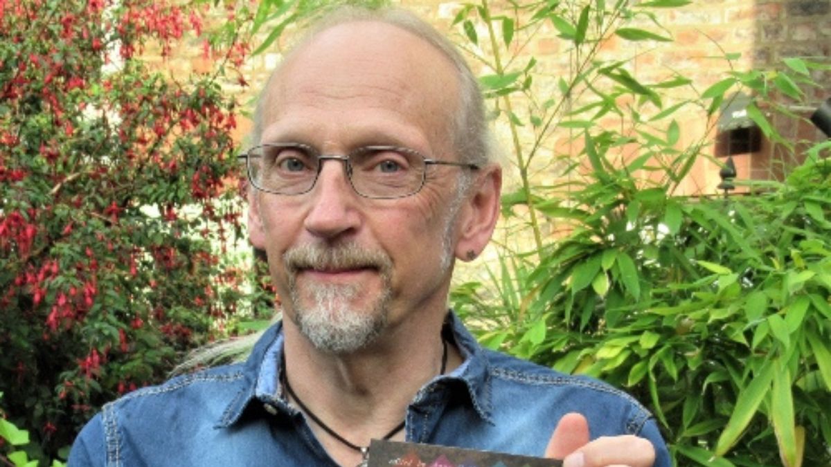 A man wearing glasses and blue shirt smiles while stood in front of green bushes.