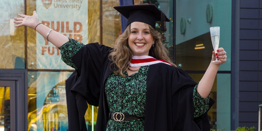 Smiling female graduate in cap and gown poses with arms outstretched.