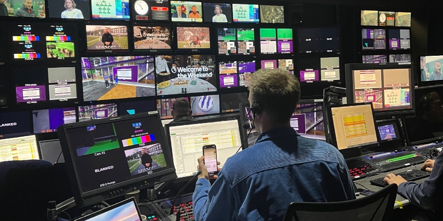 A director in a blue shirt sits at a media production desk, watching a number of different screens.