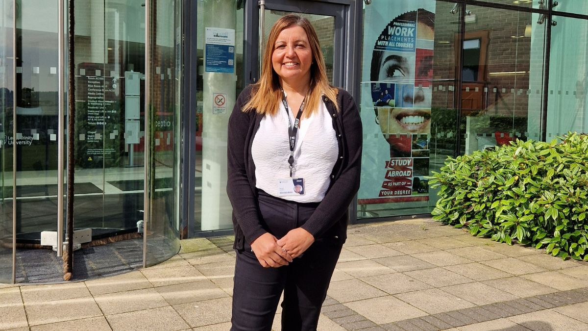 A woman wearing a white top a black cardigan and black trousers smiles for a picture in front of a building.