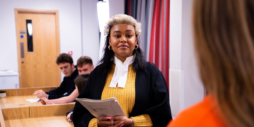 A student in solicitors court dress and yellow jumper, holding papers.