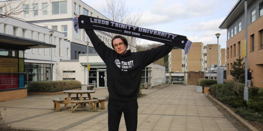 Man with dark hair and glasses stands in front of building holding Leeds Trinity University scarf above his head.