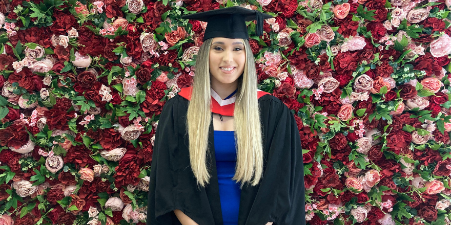 A girl with blonde hair wearing a black graduation cap and a black gown over a blue dress stands in front of a wall of red flowers.