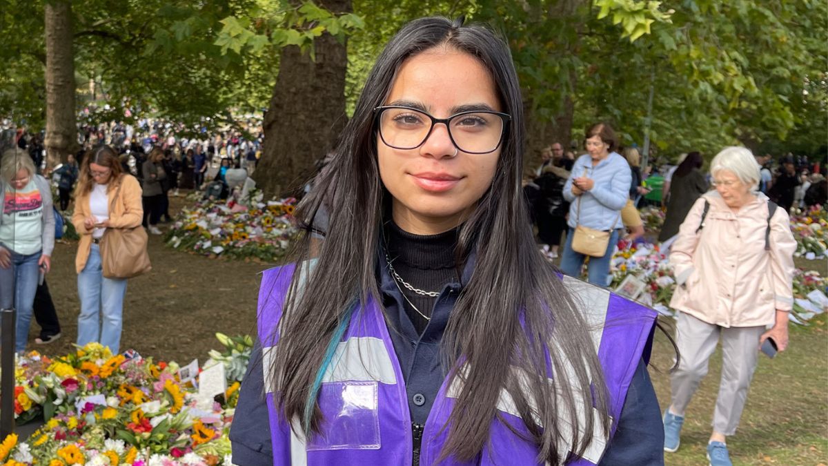 A girl with dark hair and glasses in a purple vest smiles for a picture in front of trees and flowers.