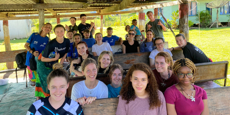 Large group of female and male students sit outside under wooden hut.