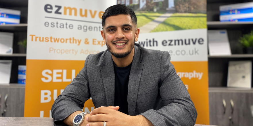 Male sits at desk smiling in front of Ezmuve sign.