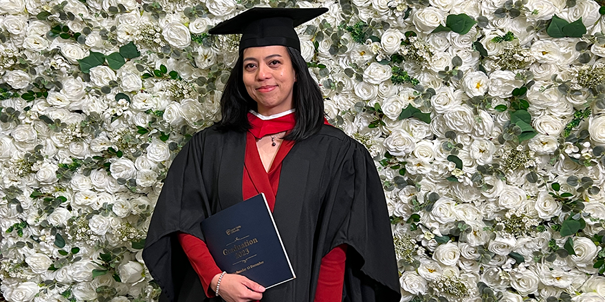 A Filipino woman in Graduation cap and gown posing in front of a wall covered in white flowers..