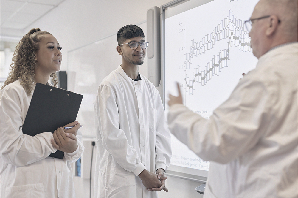 Two students in lab coats speaking to a Biomedical Science lecturer.